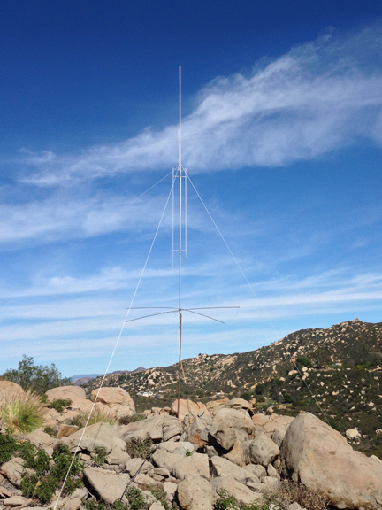 Figure 1 — The GAP Titan DX in the field — the elevated center feed partway up the mast, the four rigid counterpoise rods splayed at the base, and guy ropes carrying the mechanical load the radiator itself do…