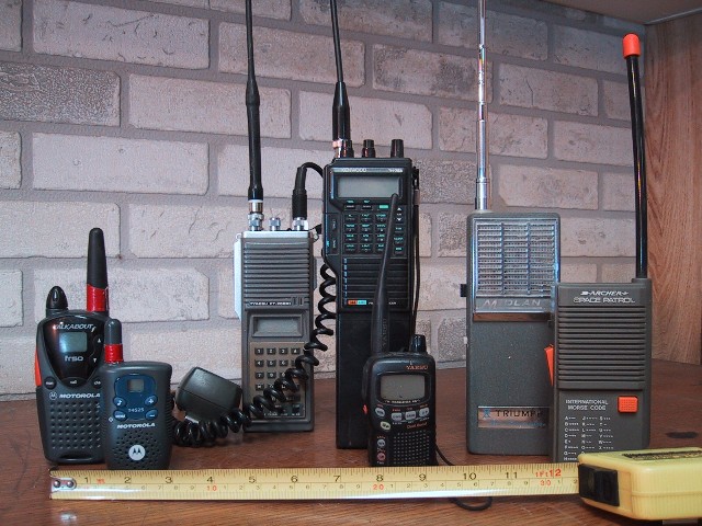 Figure 1 — A lineup of handheld radios and their factory-issue whip and helical antennas — the "before" state every commercial and DIY upgrade in this volume improves on. Photo: File:Recreational Walkie Talki…